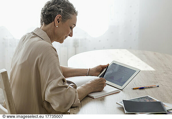 Woman holding digital tablet with data and taking notes at table at home