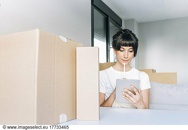 Woman holding digitized pen using tablet PC sitting with cardboard boxes at table in living room