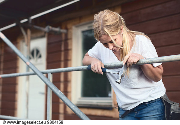 Woman holding hammer while leaning on scaffolding outside house being renovated