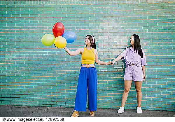 Woman holding hand of friend with multi colored balloons in front of turquoise wall