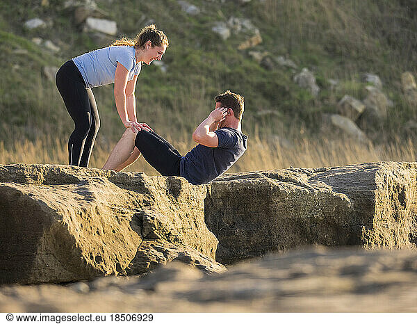 Woman holding legs of man doing crunches outdoors