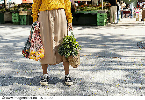 Woman holding mesh bags with fruits and vegetables standing on road at farmer's market