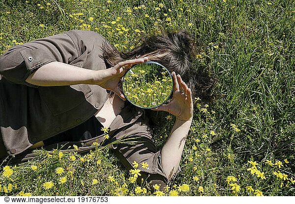 Woman holding mirror over face and lying on grass in field