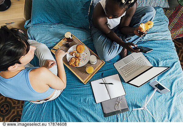 Woman holding mobile phone having breakfast with girlfriend on bed