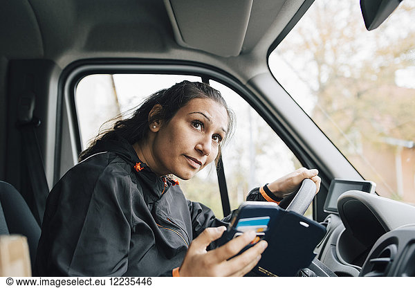 Woman holding mobile phone looking away while driving in delivery van