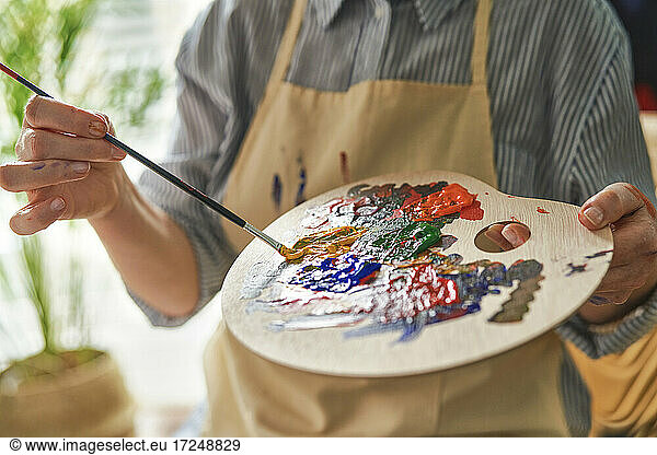 Woman holding palette while mixing colors through paintbrush at home