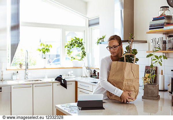 Woman holding paper bag while looking at digital tablet in kitchen