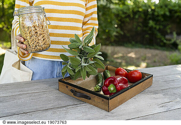 Woman holding pasta jar near wooden table with vegetable tray in garden