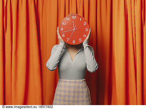 Woman holding red clock over face in front of orange curtain