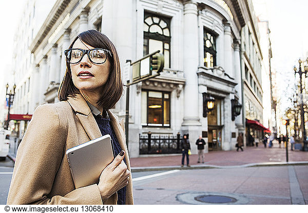 Woman holding tablet looking away while standing at city street