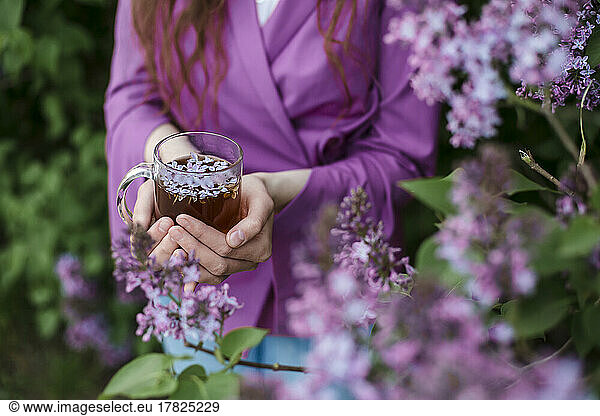 Woman holding tea cup with lilac petals by flowering plant
