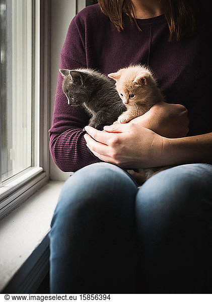 Woman holding two adorable kittens in her arms next to a window.