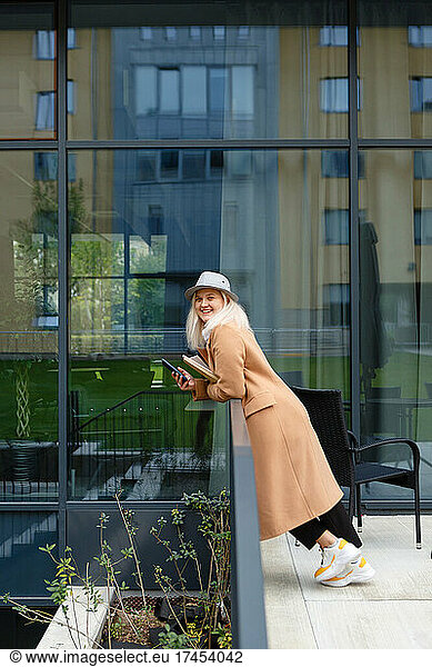 woman in a beige coat and hat holding a notebook and a smartphone