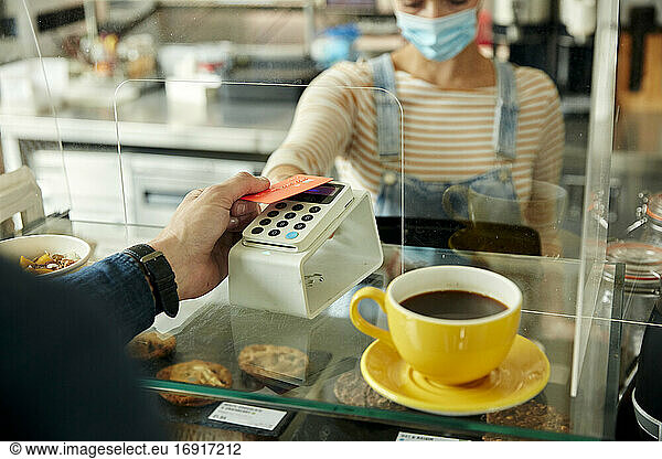 Woman in a face mask behind cafe safety screen  offering a contactless payment terminal to customer