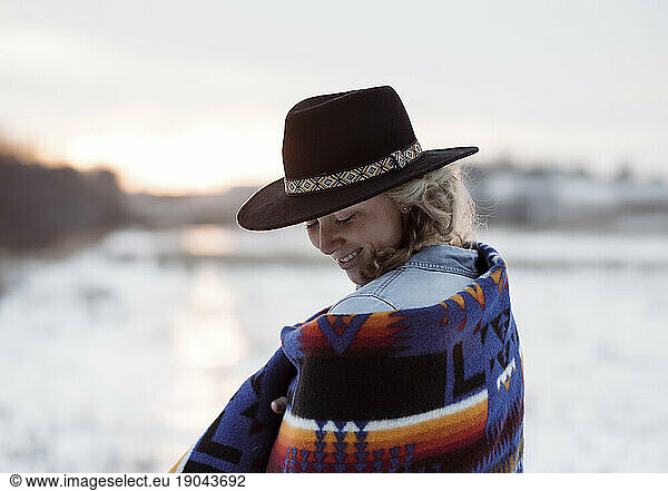 woman in a hat wrapped in a blanket looking down smiling at sunset