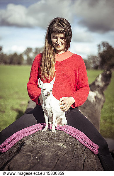 Woman in bright red jumper sits on log with small white dog on farm