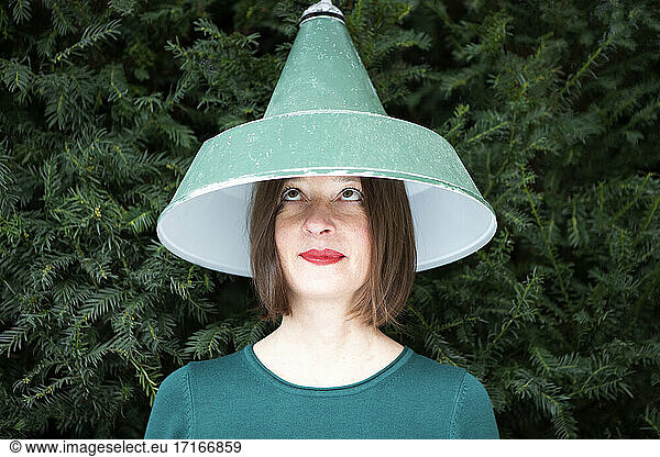 Woman in brown hair with lamp shade on head looking up against plant