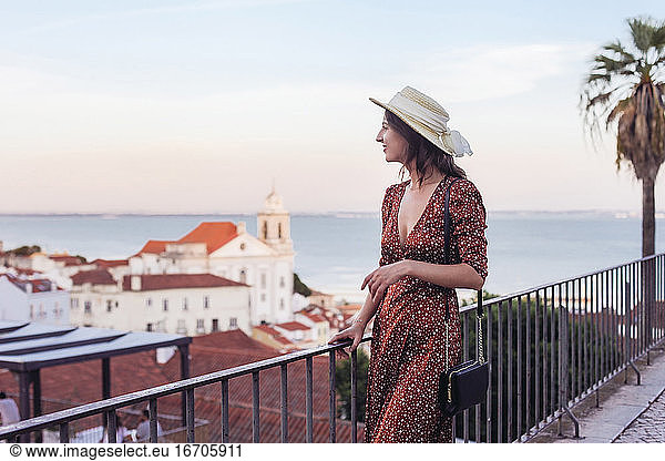Woman in dress and a straw hat looking over rooftop view of Lisbon