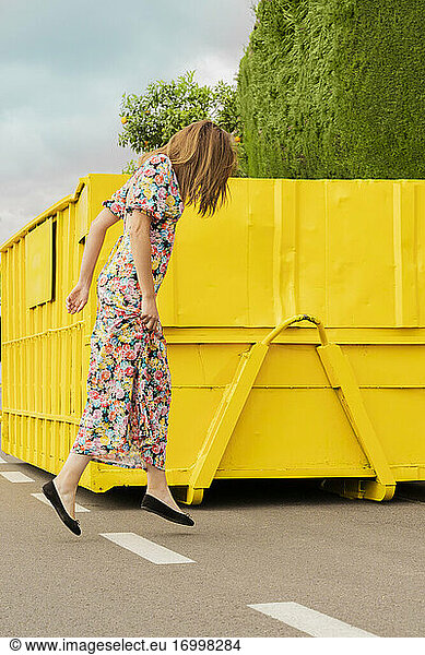 Woman in flower dress  jumping in the street in front of yellow container