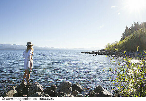 Woman in hat stands on shoreline and looks across lake
