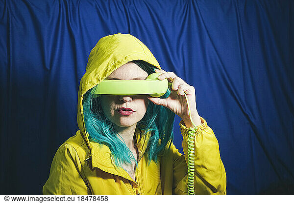 Woman in hooded shirt covering eyes with telephone receiver in front of blue backdrop