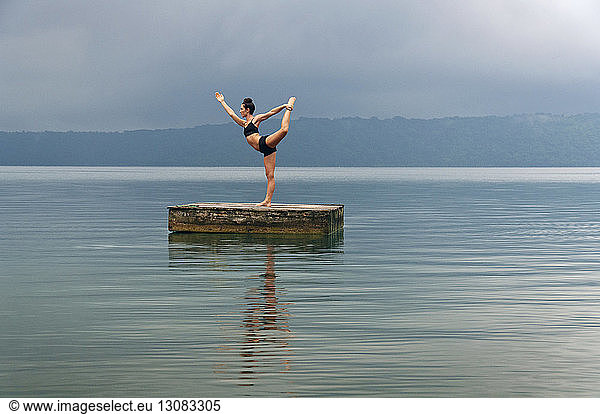 Woman in King Dancer Pose doing yoga on floating platform amidst lake against sky