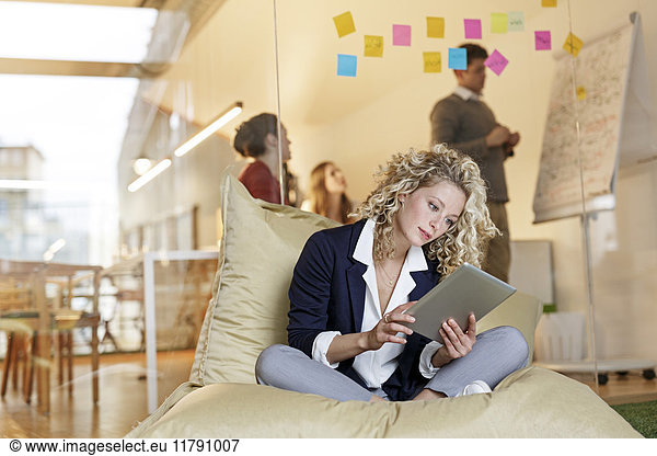 Woman in office using tablet in bean bag with meeting in background