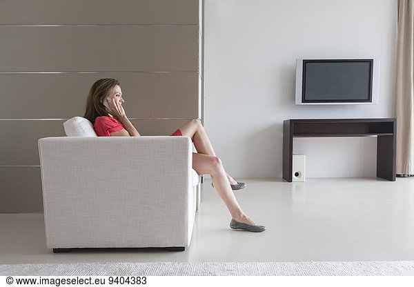 Woman in red dress sitting in armchair and talking on phone in modern living room
