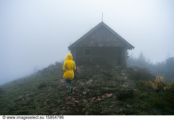 Woman in yellow coat walking through fog towards a cabin