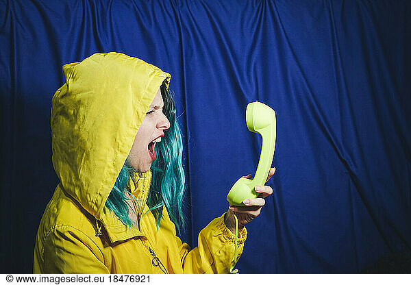 Woman in yellow hooded shirt shouting into telephone in front of blue backdrop