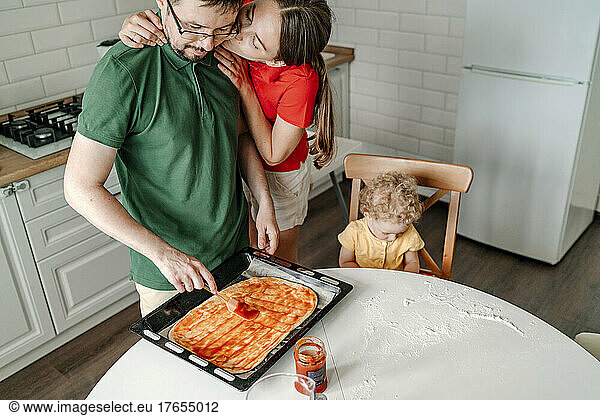 Woman kissing man preparing pizza by daughter sitting on chair in kitchen