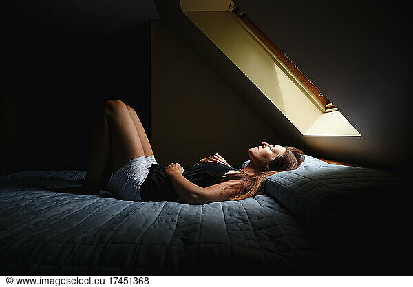Woman laying on bed with eyes closed under a window in a dark room.
