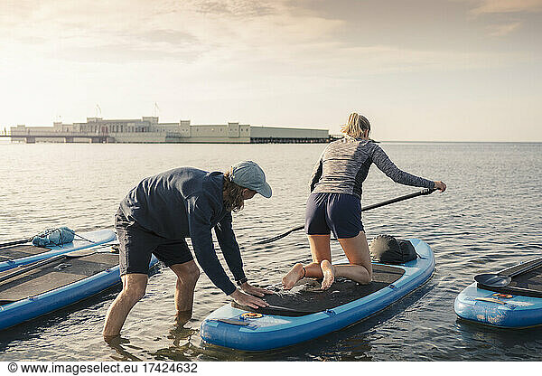Woman learning paddleboarding from male instructor in sea during sunset