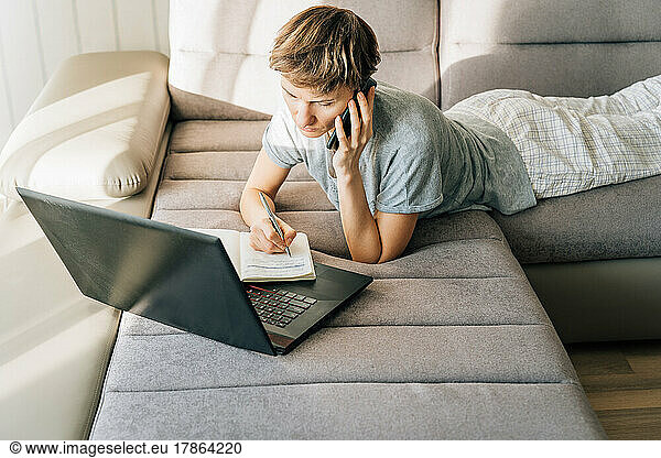 Woman lies on couch  talking on the phone and taking notes