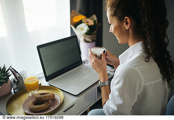 Woman looking at he laptop and smiling while holding a cup of co