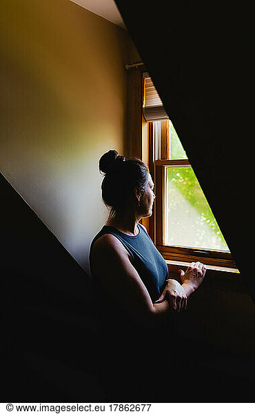 Woman looking out of a dormer window in a dark room in the daytime.