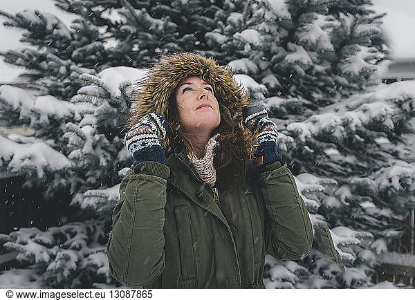 Woman looking up while wearing fur coat against trees during snowfall