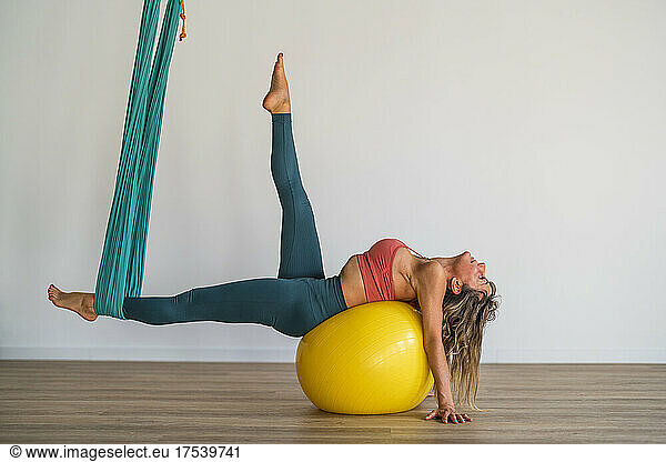 Woman lying on fitness ball stretching with aerial silk in yoga studio