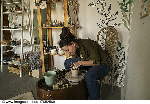 Woman making clay pot on pottery wheel sitting on chair at workshop