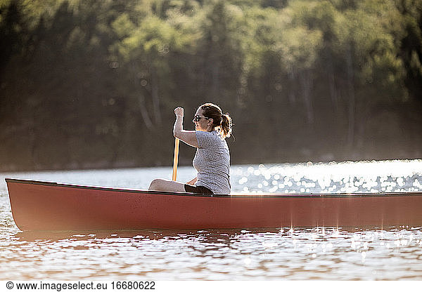 Woman paddles red canoe across lake in sunshine in Maine woods
