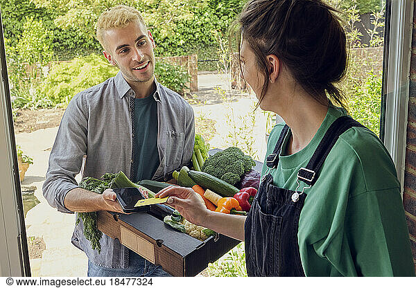 Woman paying and talking to delivery person with fresh vegetables near doorway