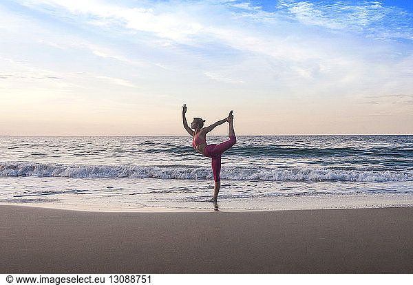 Woman performing king dancer pose at beach against sky at sunset