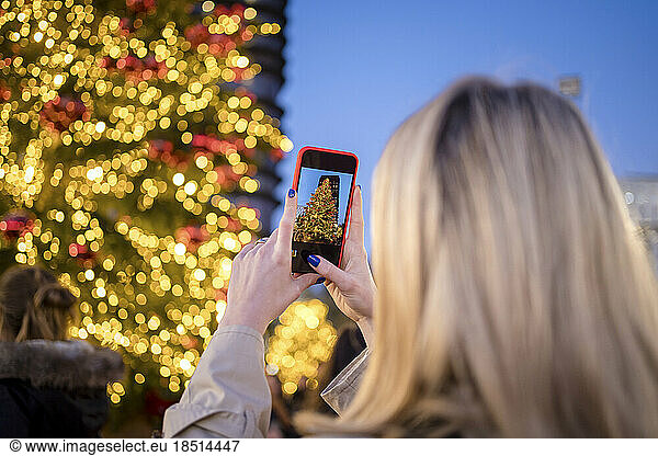 Woman photographing illuminated Christmas tree through smart phone at night