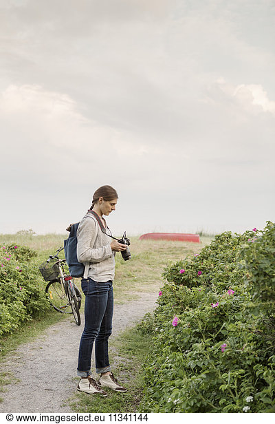 Woman photographing plants while standing on dirt road against sky