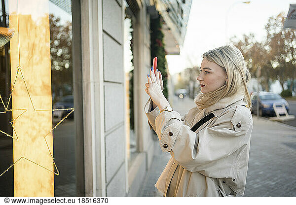 Woman photographing store window through smart phone on sidewalk