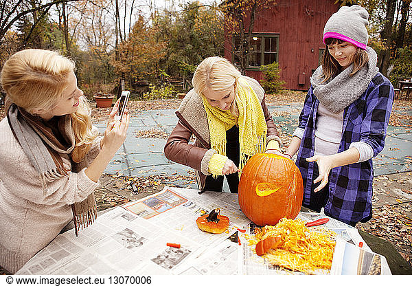 Woman photographing while friends carving Halloween pumpkin at table