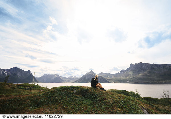 Woman photographing while sitting on mountain by lake