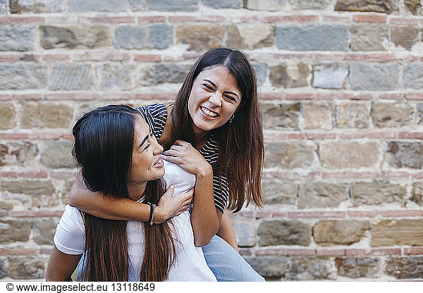 Woman piggybacking happy friend while standing by wall