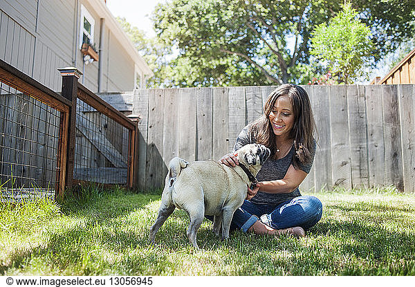 Woman playing with pug while sitting on grassy field in backyard
