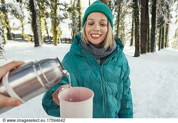 Woman pouring water in mug being held by friend at winter forest